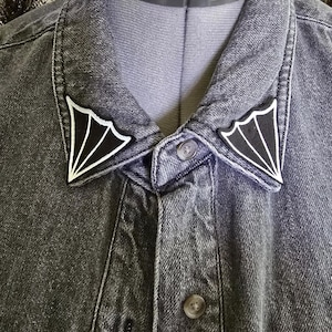 May include: Close-up of a dark gray denim jacket with decorative black and white wing-shaped patches on the collar. The jacket is buttoned up, and a gray choker necklace is visible.