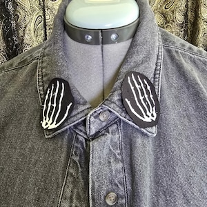 May include: A gray denim jacket with two black patches featuring white bone hand designs on the collar. The jacket is buttoned up and displayed on a mannequin.