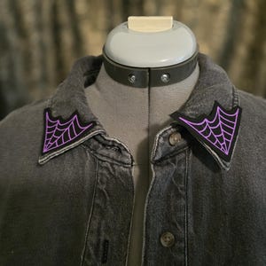 Può includere: Giacca di jeans grigia con due toppe a forma di ragnatela nere e viola sul colletto. Le toppe sono triangolari e presentano un design a ragnatela dettagliato. La giacca è abbottonata e mostrata su un manichino.