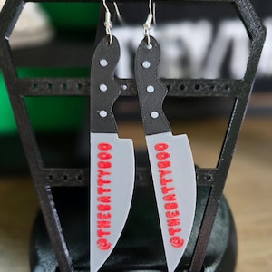 May include: A pair of novelty earrings shaped like miniature knives. The blades are gray with the text "@THEBATTYBOO" in red. The handles are black with white dot details. The earrings hang from silver-colored hooks.