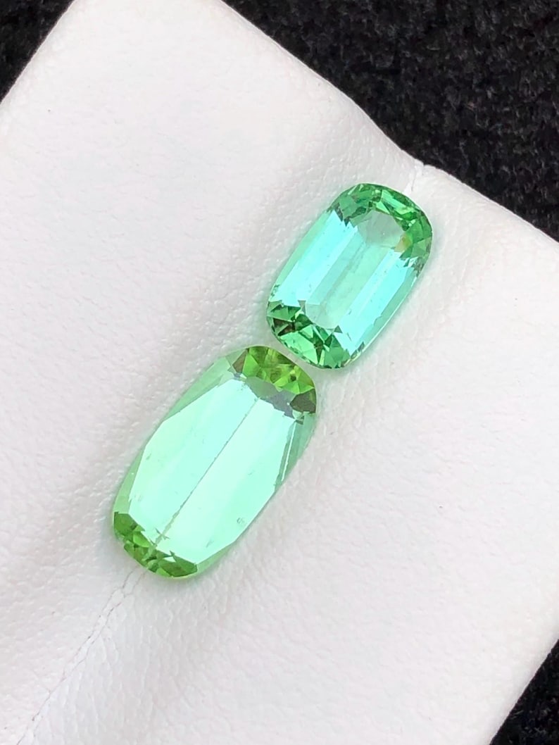 May include: Two rectangular-cut gemstones with a vibrant, light green colour. The gems are displayed on a white surface, showcasing their clarity and brilliance. The stones have a slight blue undertone, adding depth.