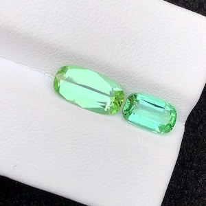 May include: Two light green, rectangular gemstones with faceted edges. The gems are displayed on a white surface, with a dark background. The gemstones have a bright, translucent appearance.