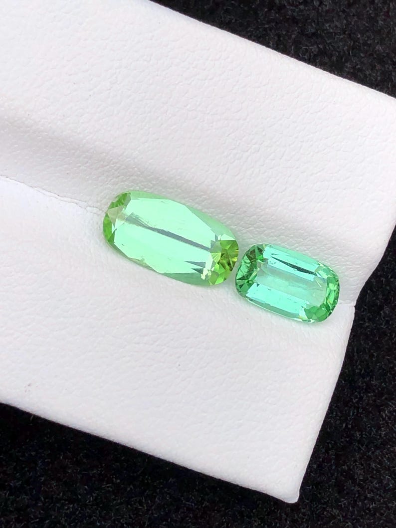 May include: Two light green, rectangular gemstones with faceted edges. The gems are displayed on a white surface, with a dark background. The gemstones have a bright, translucent appearance.