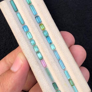May include: A collection of twenty-two loose, faceted gemstones arranged in two parallel rows on a white display pad. The stones are primarily rectangular and baguette-cut, featuring a variety of colors including shades of blue, green, and pink.