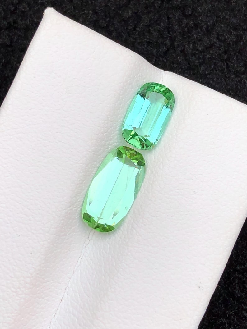 May include: Two light green, rectangular gemstones with rounded edges. The gems have a slight blue hue and are set against a white surface. The gems are faceted, reflecting light.