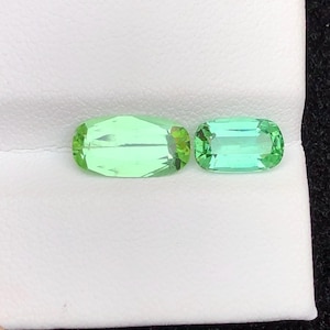 May include: Two light green gemstones with a rectangular shape and rounded edges. The gemstones are displayed on a white surface. The larger gemstone is approximately 2.5 cm in length, and the smaller gemstone is about 2 cm long. The gemstones have a slight sparkle.