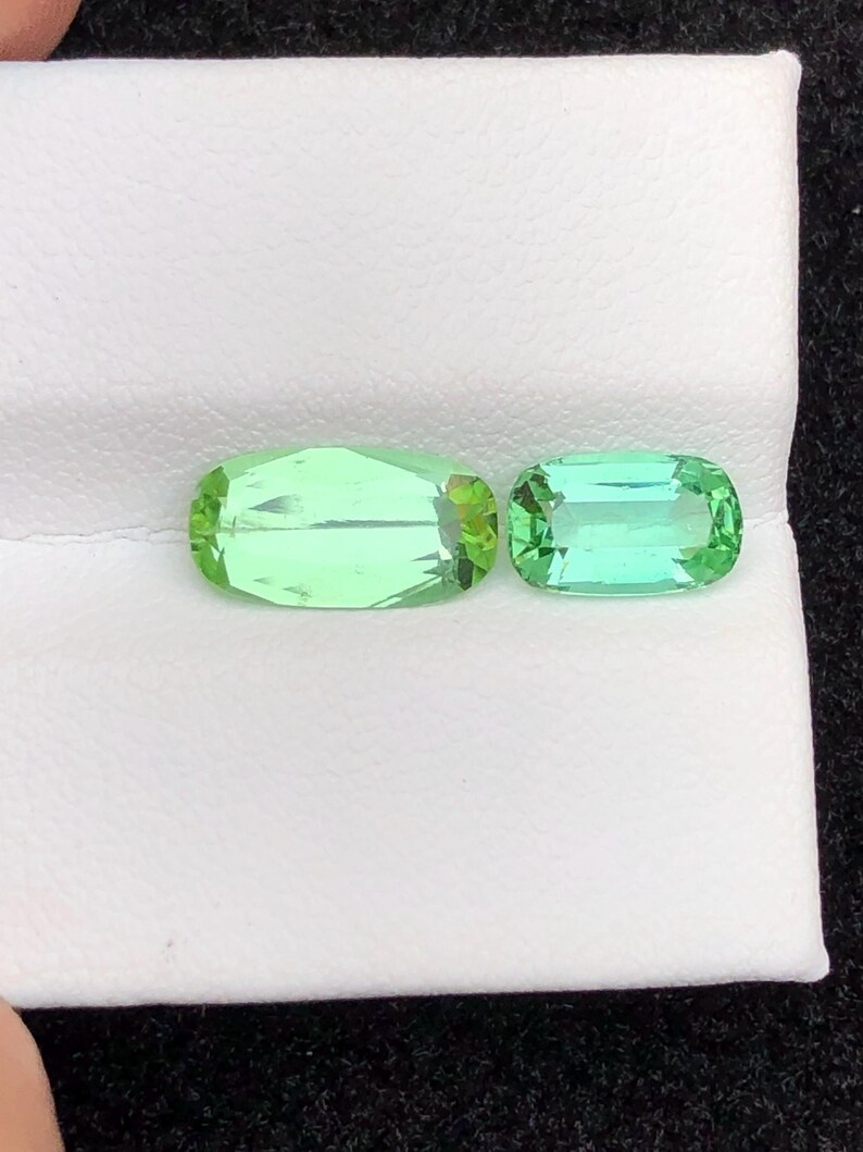 May include: Two light green gemstones with a rectangular shape and rounded edges. The gemstones are displayed on a white surface. The larger gemstone is approximately 2.5 cm in length, and the smaller gemstone is about 2 cm long. The gemstones have a slight sparkle.