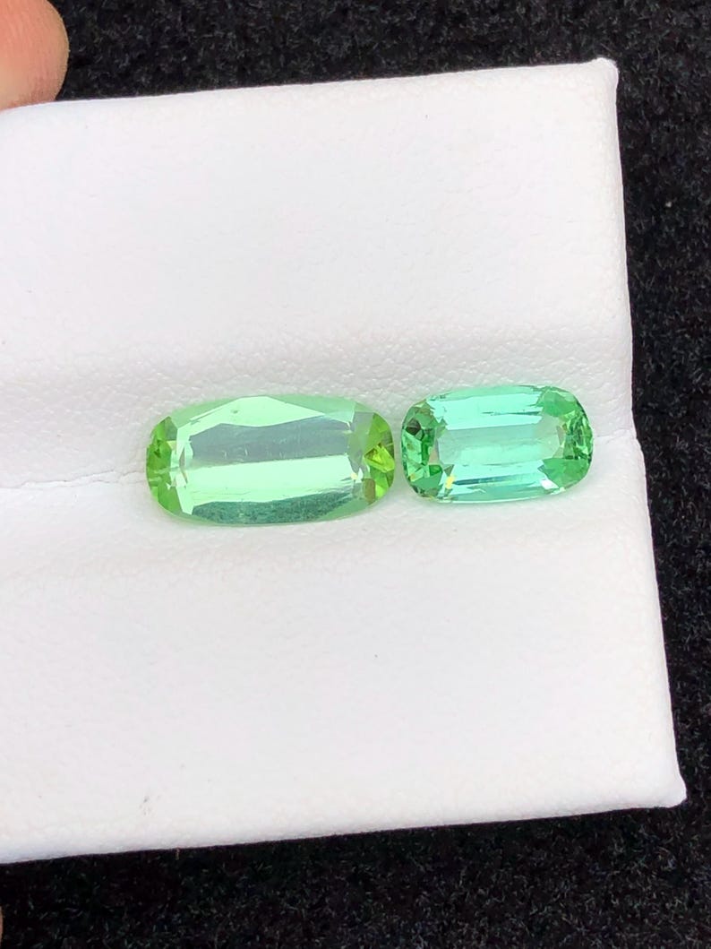May include: Two light green, rectangular gemstones with rounded corners are displayed on a white surface. The gems have a transparent appearance, with visible facets. The larger gem is approximately 2.5 cm long, and the smaller gem is about 2 cm long.