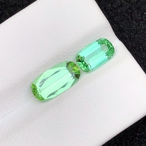 May include: Two light green, rectangular gemstones with faceted edges are displayed on a white surface. The gems have a vibrant, transparent appearance, suggesting they are cut and polished. The background is a dark, textured fabric.