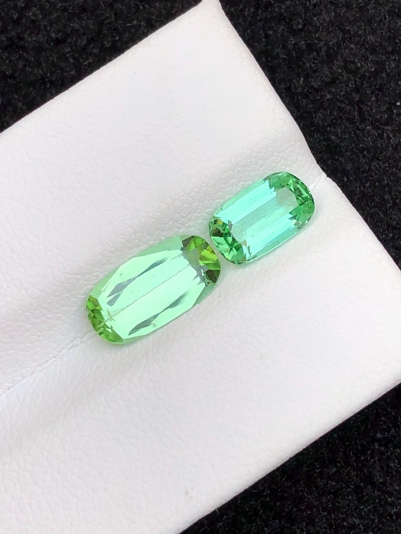 May include: Two light green, rectangular gemstones with faceted edges are displayed on a white surface. The gems have a vibrant, transparent appearance, suggesting they are cut and polished. The background is a dark, textured fabric.