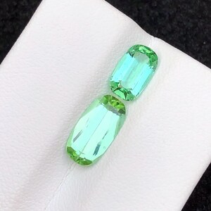 May include: Two rectangular, faceted gemstones with a light green and blue hue. The gems are displayed on a white surface. The stones have a polished, reflective surface, showcasing their clarity and colour. These gems are likely for jewellery making.