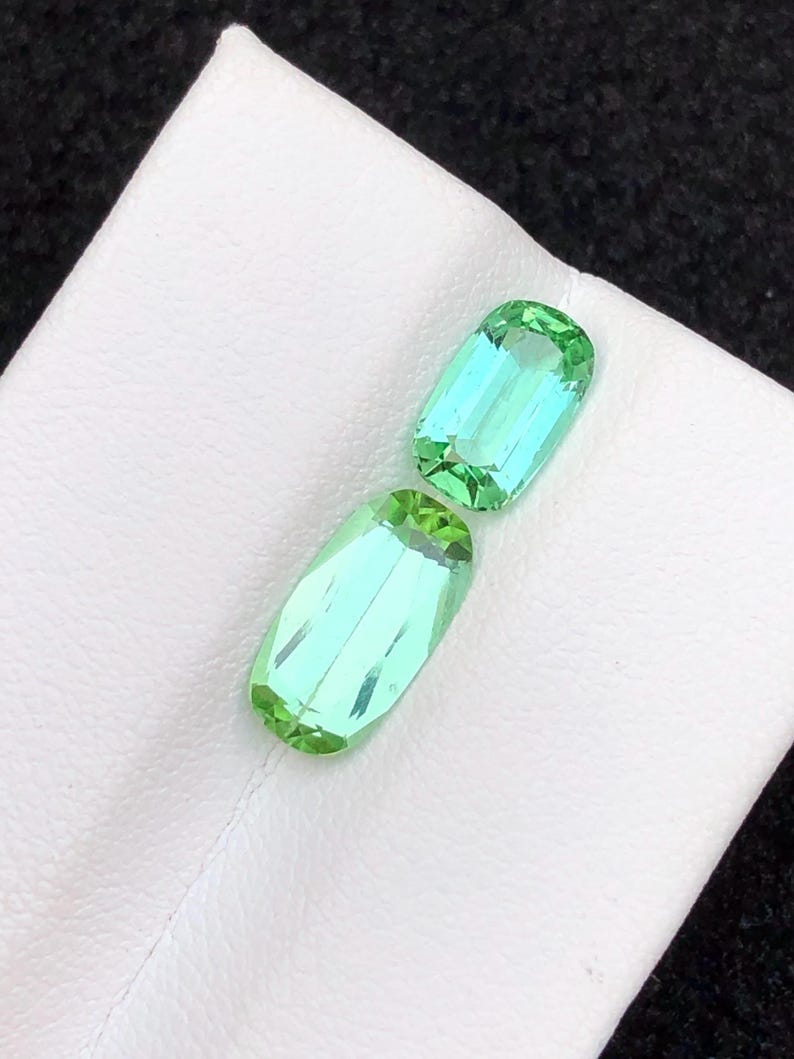 May include: Two rectangular, faceted gemstones with a light green and blue hue. The gems are displayed on a white surface. The stones have a polished, reflective surface, showcasing their clarity and colour. These gems are likely for jewellery making.