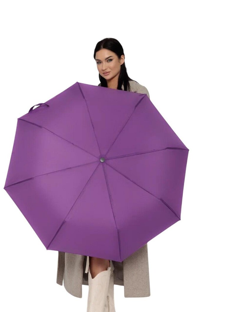 Peut inclure: Une femme tient un parapluie violet au-dessus de sa t&ecirc;te. Le parapluie est ouvert et la femme regarde la cam&eacute;ra.