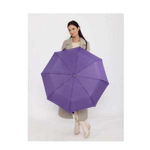 Puede incluir: Un paraguas morado vibrante, completamente abierto, sostenido por una persona. El color del paraguas es un tono profundo y saturado. La persona lleva un abrigo y botas de tonos neutros, creando un contraste con el paraguas.