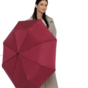 Peut inclure: Une femme tenant un parapluie bordeaux. Le parapluie est ferm&eacute; et a une poign&eacute;e noire.