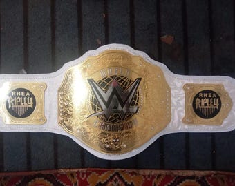 Puede incluir: Un cinturón de campeonato de lucha libre blanco y dorado con las palabras "World Championship" y un logotipo "W" en blanco y negro. El cinturón tiene dos placas laterales con el nombre "Rhea Ripley" en blanco y negro.