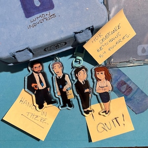 May include: Four clear acrylic keychains featuring cartoon characters from the TV show Severance. The characters are Helly R, Mark S, Irving, and Petey. The keychains are hanging from a blue background with yellow sticky notes that say "MDR Severance Keychains By Filworks", "Hang in there", "Quit!" and "Lumon Industries".