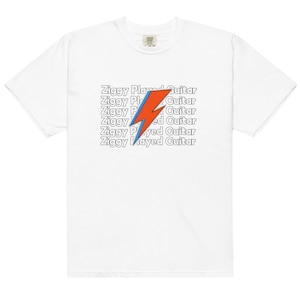 May include: White t-shirt featuring the text "Ziggy Played Guitar" stacked vertically with a red and blue lightning bolt graphic. The shirt has a crew neck and short sleeves.