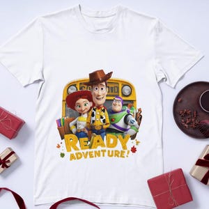 Puede incluir: Una camiseta blanca de manga corta con un estampado gráfico de Woody, Jessie y Buzz Lightyear de Toy Story frente a un autobús escolar amarillo, con el texto READY ADVENTURE en letras amarillas, rodeado de pequeños iconos de hojas de otoño.