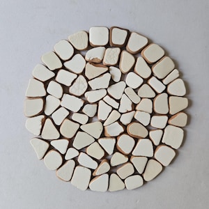 May include: Circular trivet composed of off-white and tan ceramic tiles arranged in a mosaic design. The tiles are of different shapes and sizes, creating a textured surface. The trivet is designed to protect surfaces from heat.