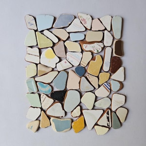 Può includere: Una collezione di frammenti di ceramica assortiti disposti in un motivo a mosaico rettangolare. I pezzi presentano vari colori tra cui azzurro, giallo, bianco, crema e marrone, con alcuni frammenti che mostrano motivi floreali e linee geometriche.
