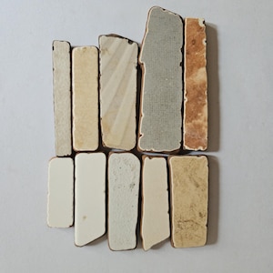 Può includere: Un assortimento di piastrelle rettangolari in ceramica in vari colori e texture. Le piastrelle includono tonalità di crema, beige, grigio e marrone, alcune con motivi a righe o macchiati. I bordi sono ruvidi, conferendo loro un aspetto rustico.