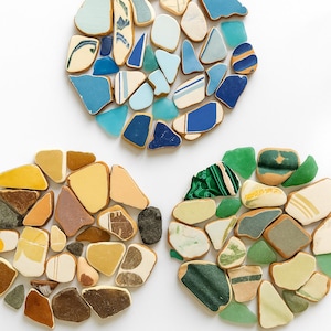 以下が含まれることがあります： 青、茶色、黄色、緑の色合いのシーグラスの破片を円形に3つ配置。破片のサイズと形状は様々で、模様や質感のあるものもある。背景は無地の白。