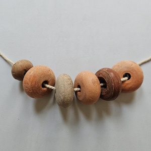 May include: A necklace featuring six circular, earthy-toned beads strung on a light-colored cord. The beads vary in color from light tan and beige to shades of terracotta and dark brown, each displaying a textured, matte, and porous surface finish.