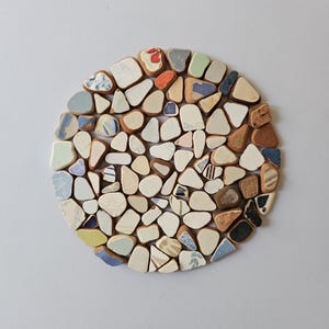Puede incluir: Un salvamanteles redondo hecho de pequeños trozos de cerámica de forma irregular en varios colores, incluyendo blanco, azul, marrón y verde. Las piezas están dispuestas muy juntas, creando un patrón similar a un mosaico. El salvamanteles está diseñado para proteger las superficies del calor.