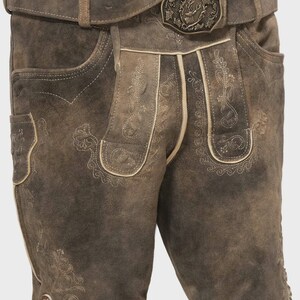 Pode incluir: Calções de couro castanho tipo lederhosen com bordados decorativos e cinto a condizer. O cinto apresenta uma fivela detalhada com um emblema de veado. Bolsos e botões decorativos.