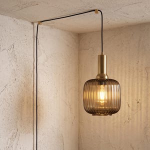 May include: A pendant light featuring a cylindrical brass fixture and a ribbed, smoke-colored glass shade. The light is suspended by a black cord that runs along the ceiling and down the wall, secured by two small brass mounting brackets.