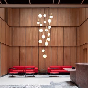 May include: A multi-pendant chandelier featuring numerous spherical white glass globes suspended by thin gold-toned metal rods at varying heights, creating a cascading vertical arrangement against a paneled wood wall in a lobby space.