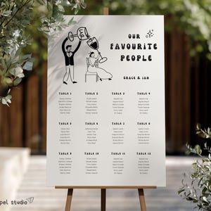 May include: A wedding seating plan displayed on a wooden easel. The white sign has a black and white illustration of a couple and the text "OUR FAVOURITE PEOPLE" and the names "GRACE & IAN". Guest names are listed under table numbers.