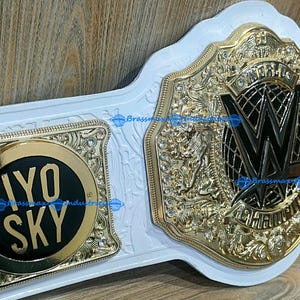 May include: A white and gold championship belt with intricate detailing. The belt features a large central plate with the WWE logo and two side plates with the text "IYO SKY". The belt is made of gold and white materials.