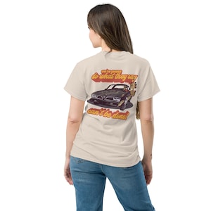 Puede incluir: Camiseta beige con un gráfico de un coche negro y el texto "we're gonna do what they say can't be done!" en rojo y amarillo. La camiseta la lleva una persona con vaqueros azules.