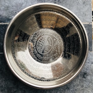Può includere: Una ciotola d'argento con calligrafia araba incisa sulla superficie. La ciotola è di forma circolare e ha una superficie riflettente. La calligrafia è in stile tradizionale e include la frase "La ilaha illa Allah" che si traduce in "Non c'è altro Dio che Dio".