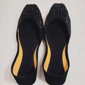 Puede incluir: Un par de bailarinas negras con punta redonda, adornadas con peque&ntilde;as piedras oscuras. El interior presenta una suela en contraste amarillo-naranja. Los zapatos se muestran sobre un fondo blanco liso, resaltando su dise&ntilde;o.