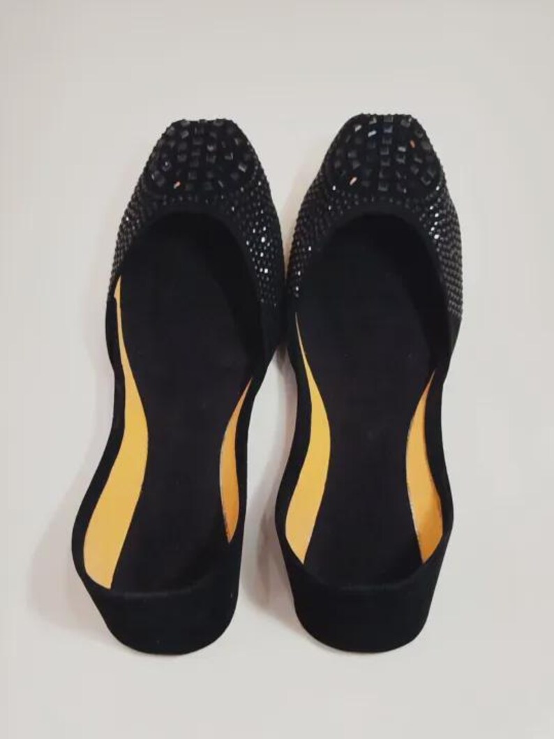 Puede incluir: Un par de bailarinas negras con punta redonda, adornadas con peque&ntilde;as piedras oscuras. El interior presenta una suela en contraste amarillo-naranja. Los zapatos se muestran sobre un fondo blanco liso, resaltando su dise&ntilde;o.