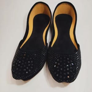 Puede incluir: Un par de bailarinas negras adornadas. Los zapatos tienen una punta redondeada y est&aacute;n cubiertos de peque&ntilde;as decoraciones oscuras de forma cuadrada. El interior es de color amarillo claro. Los zapatos se exhiben sobre una superficie blanca.