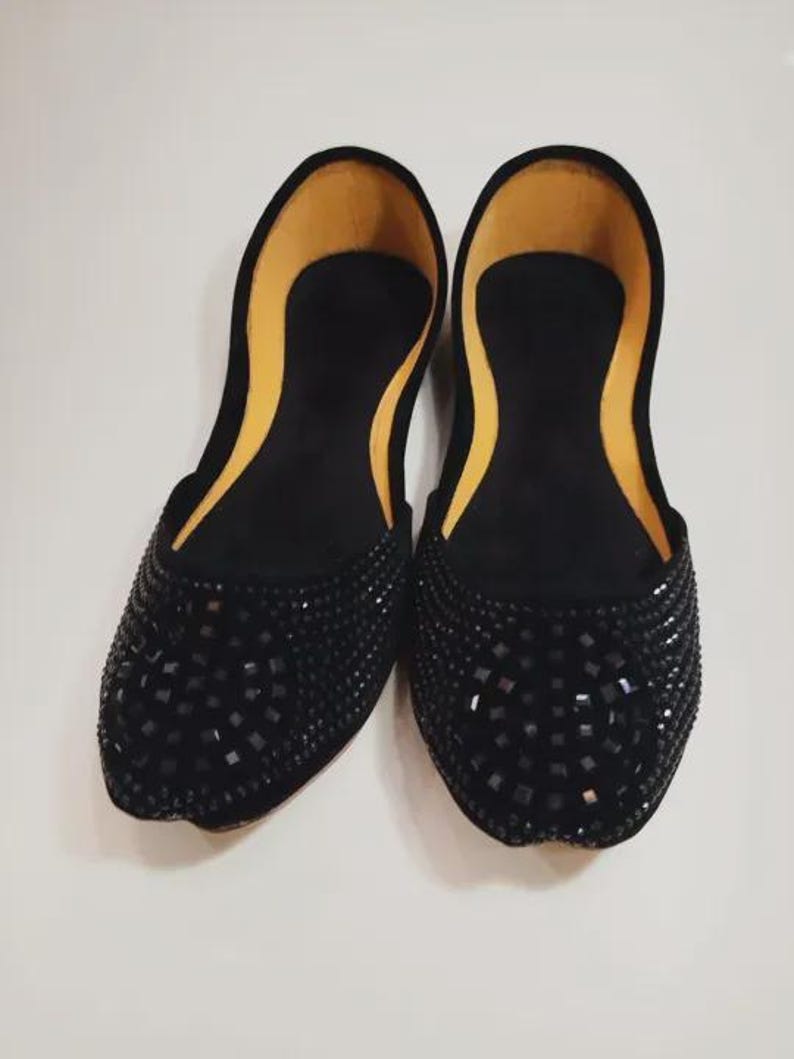 Puede incluir: Un par de bailarinas negras adornadas. Los zapatos tienen una punta redondeada y est&aacute;n cubiertos de peque&ntilde;as decoraciones oscuras de forma cuadrada. El interior es de color amarillo claro. Los zapatos se exhiben sobre una superficie blanca.