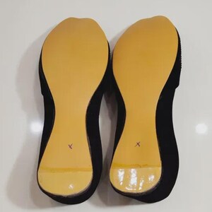 Puede incluir: Un par de bailarinas negras con suelas color canela. Las suelas tienen un acabado liso y brillante y est&aacute;n marcadas con una "X" morada. Los zapatos se muestran sobre una superficie blanca.