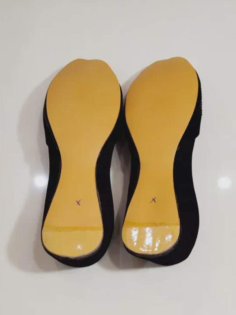 Puede incluir: Un par de bailarinas negras con suelas color canela. Las suelas tienen un acabado liso y brillante y est&aacute;n marcadas con una "X" morada. Los zapatos se muestran sobre una superficie blanca.
