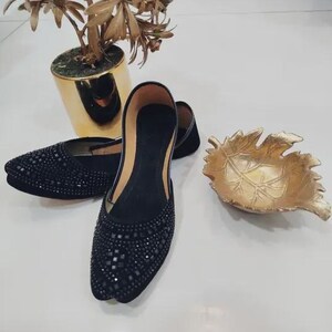 Puede incluir: Un par de bailarinas negras con punta, adornadas con peque&ntilde;as piedras oscuras. Los zapatos se exhiben junto a un cuenco decorativo dorado en forma de hoja y una planta en maceta dorada.