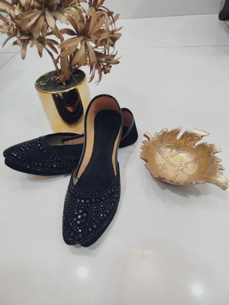 Puede incluir: Un par de bailarinas negras con punta, adornadas con peque&ntilde;as piedras oscuras. Los zapatos se exhiben junto a un cuenco decorativo dorado en forma de hoja y una planta en maceta dorada.