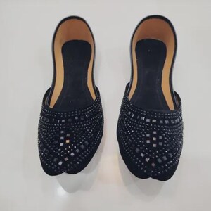 Puede incluir: Un par de zapatos negros sin cordones con un patr&oacute;n decorativo de peque&ntilde;os adornos cuadrados. Los zapatos tienen una punta redondeada y un interior de color beige.