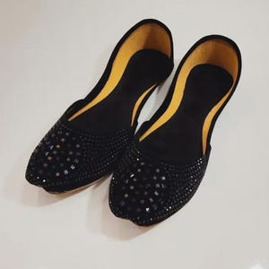 Puede incluir: Un par de bailarinas negras con punta, adornadas con peque&ntilde;os detalles cuadrados oscuros. El forro interior es de color amarillo brillante. Los zapatos se exhiben sobre una superficie blanca.