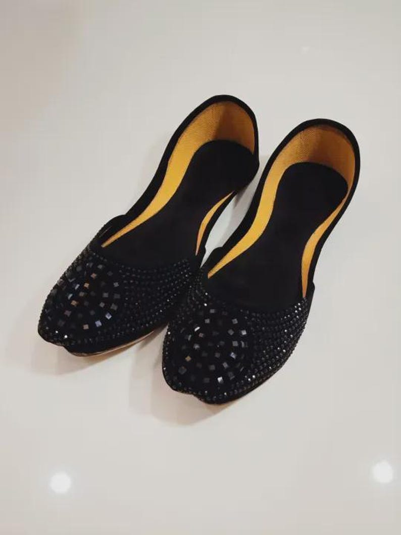 Puede incluir: Un par de bailarinas negras con punta, adornadas con peque&ntilde;os detalles cuadrados oscuros. El forro interior es de color amarillo brillante. Los zapatos se exhiben sobre una superficie blanca.