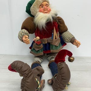 May include: A doll featuring a green hat with faux fur trim, a red velvet tunic, and brown sleeves with a plaid pattern. The doll has a long white beard, a brown belt with pockets containing a blue pencil and ruler, and brown curled shoes with bells.
