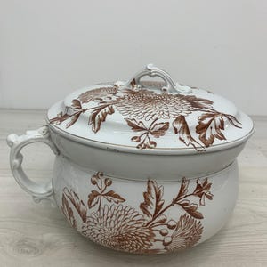 Może przedstawiać: Biały ceramiczny nocnik z pasującą pokrywką, ozdobiony brązowym wzorem kwiatowym przedstawiającym chryzantemy i liście. Naczynie posiada pojedynczy ozdobny uchwyt boczny oraz dekoracyjny uchwyt na pokrywce, wykończony błyszczącą glazurą.