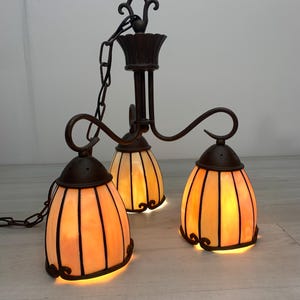 Op de afbeelding: Een hangende kroonluchter met drie lampen, voorzien van een donkerbruin metalen frame met gebogen armen en een ketting. Elke lamp heeft een klokvormige kap van warme, amberkleurige glaspanelen met een donkere metalen rand.